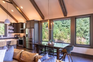 Een moderne keuken met roestvrijstalen apparatuur, een eethoek met tafel en stoelen, en grote ramen met uitzicht op groen.