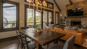 Um quarto espaçoso com uma grande mesa de jantar de madeira, assentos confortáveis, uma lareira de pedra e janelas amplas que oferecem vista para o exterior.