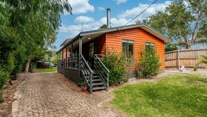A wooden cabin with a porch and a chimney, surrounded by greenery and a paved path. - WAVELENGTH - Free Wifi and Pet Friendly (outside only) (Inverloch)