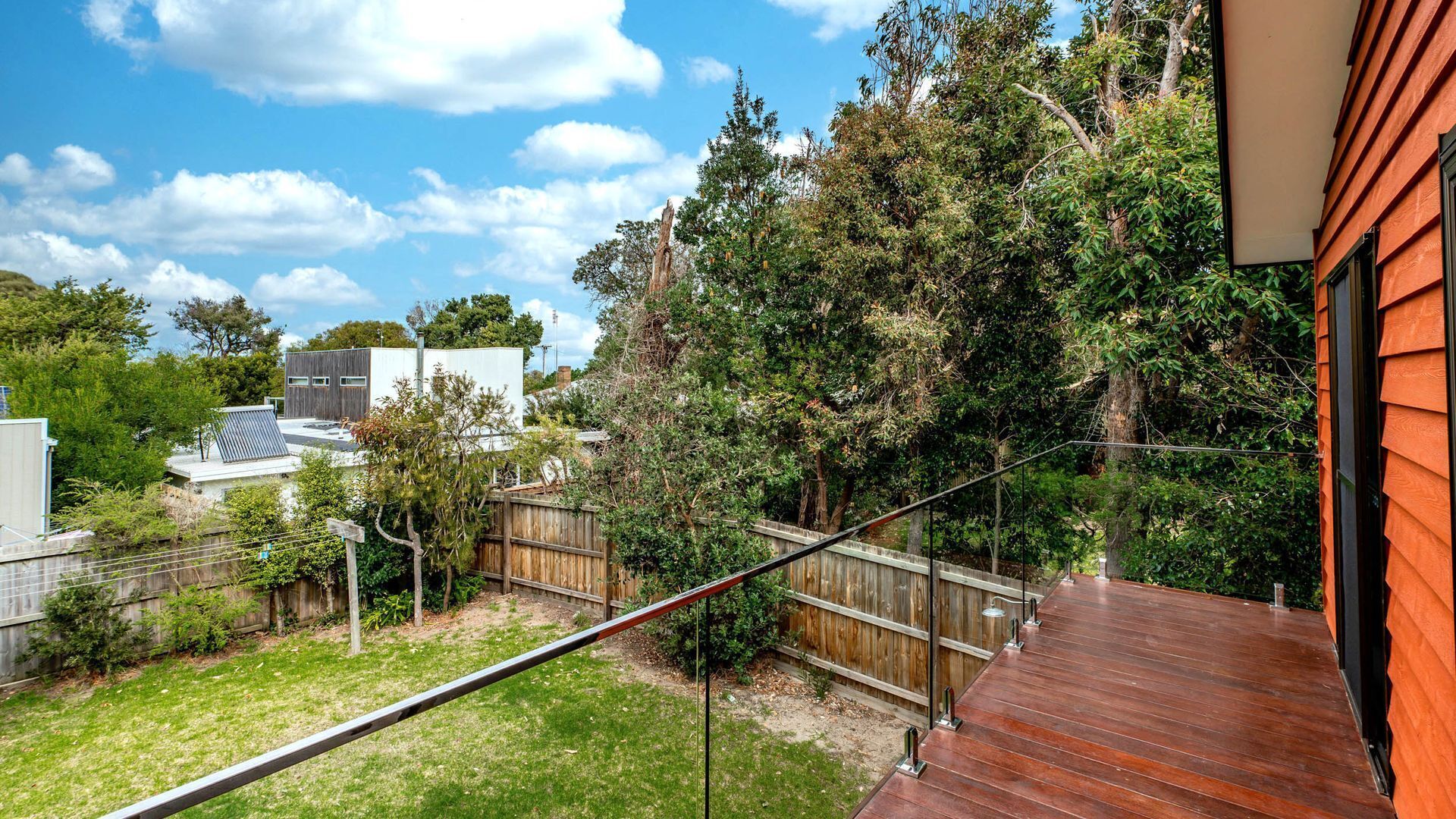 Un jardin à l’arrière d’une maison, avec une terrasse en bois, un escalier en métal et une zone délimitée par une clôture.