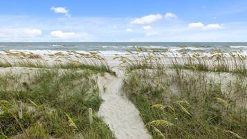 Un sentier de sable qui longe la plage et mène à l’océan, avec des dunes et de l’herbe.