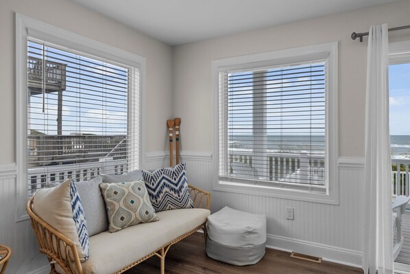 A cozy room with a wicker sofa, two patterned cushions, and a matching ottoman. Large windows with white blinds offer a view of the ocean and a wooden deck.