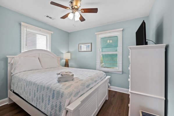 Un dormitorio con cama, ventilador de techo y dos ventanas con persianas.