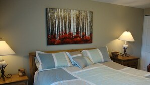 A neatly made bed with a white and blue bedspread, two bedside tables with lamps, a small wooden nightstand, and a painting of a forest on the wall. - 2King Master Suites, Walk to golf, Outside heated pool (Cedar)