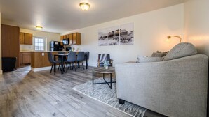 Moderni huoneisto, jossa on keittiö, ruokailutila ja olohuone. Huoneistossa on puukalusteet, ruokapöytä tuoleineen, harmaa sohva ja matto.