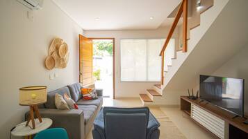 Un salon moderne avec un canapé, un fauteuil bleu, un téléviseur et un escalier menant à l’étage supérieur.