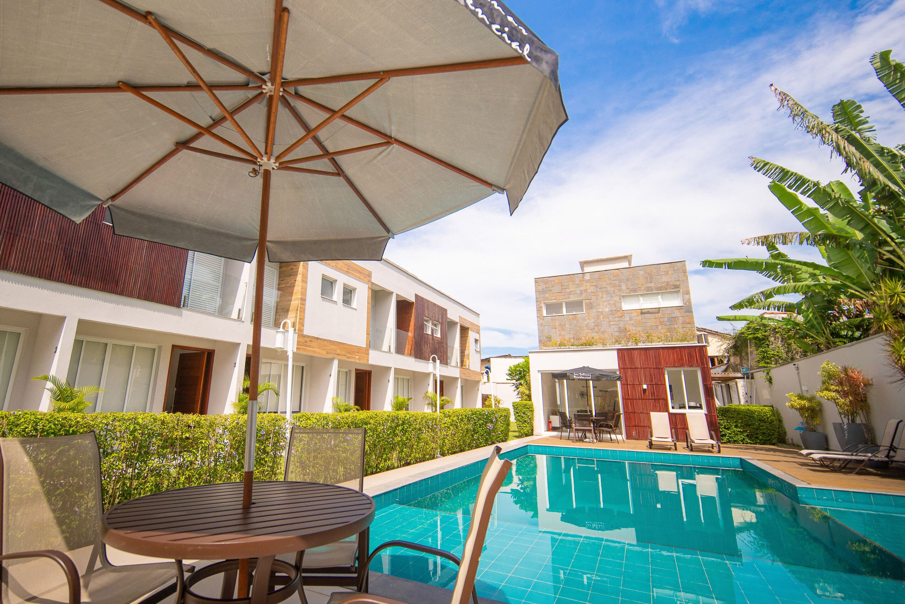 A swimming pool area with a table and chairs under a large umbrella, surrounded by buildings and greenery.