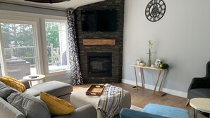 A living room with a stone fireplace, a grey sofa, a blue armchair, and a small side table with decorative items. - Waterview 5 bedroom home.  Sleeps 10 (Cornwall)
