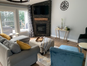Uma sala de estar com lareira de pedra, um sofá cinza, uma poltrona azul e uma mesinha lateral com objetos decorativos.
