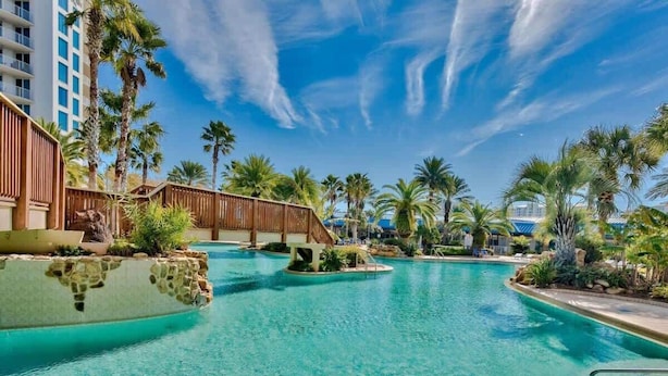 Un espace piscine aménagé dans un complexe hôtelier, comprenant plusieurs éléments aquatiques, des palmiers et un pont en bois.