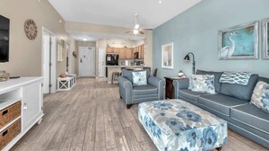 Un amplio espacio de sala con un sofá, reposapiés y televisor. Se ve una cocina al fondo.