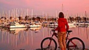 Eine Person steht neben einem Fahrrad an einer Marina, im Hintergrund sind Boote zu sehen.