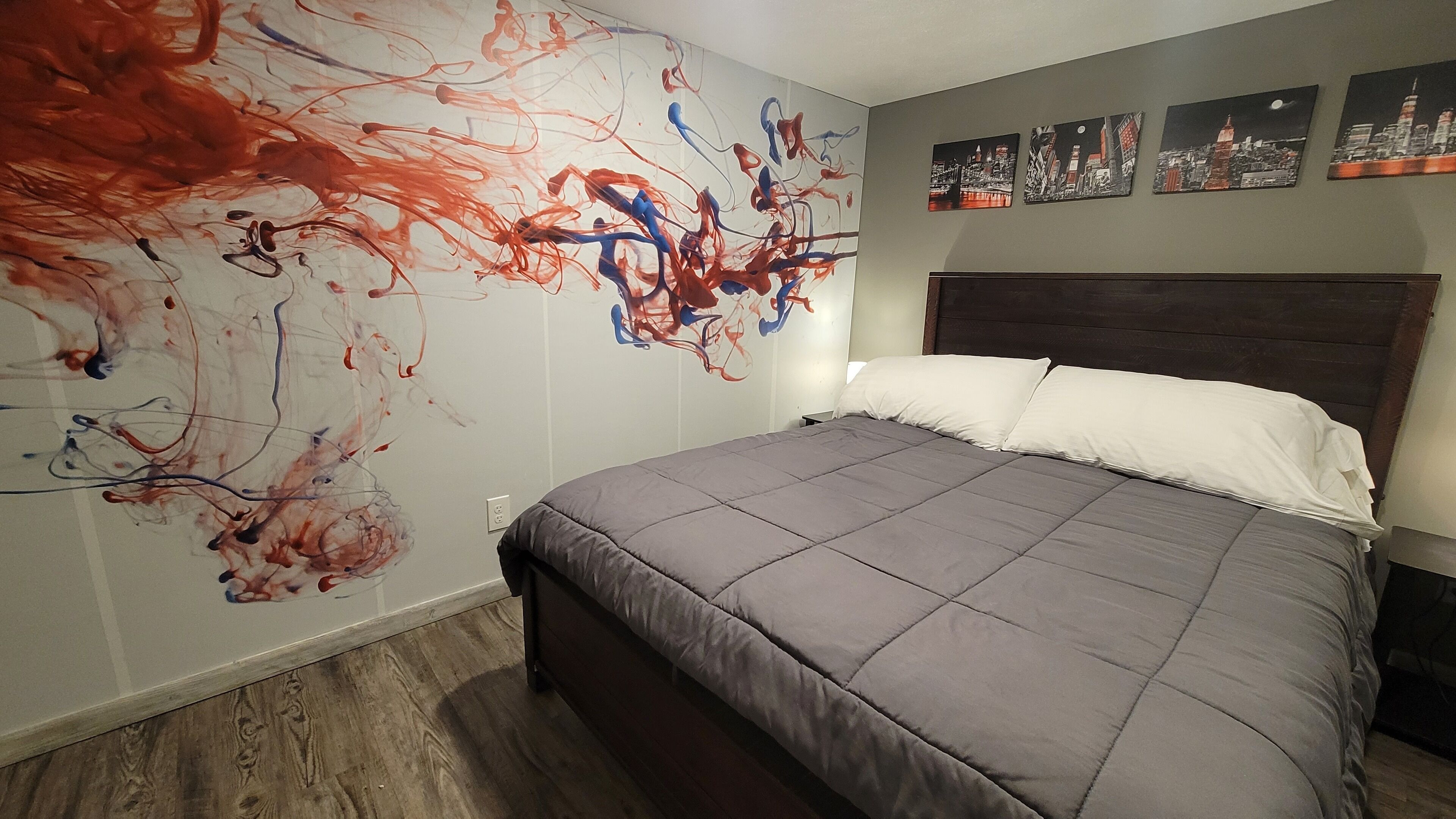 Een slaapkamer met een groot abstract schilderij aan de muur, een bed met grijze beddengoed en stadsgezichten erboven.