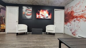 Une pièce avec deux chaises blanches, un pouf gris et une table en bois. On y trouve deux portes blanches et un grand tableau abstrait sur le mur de droite.
