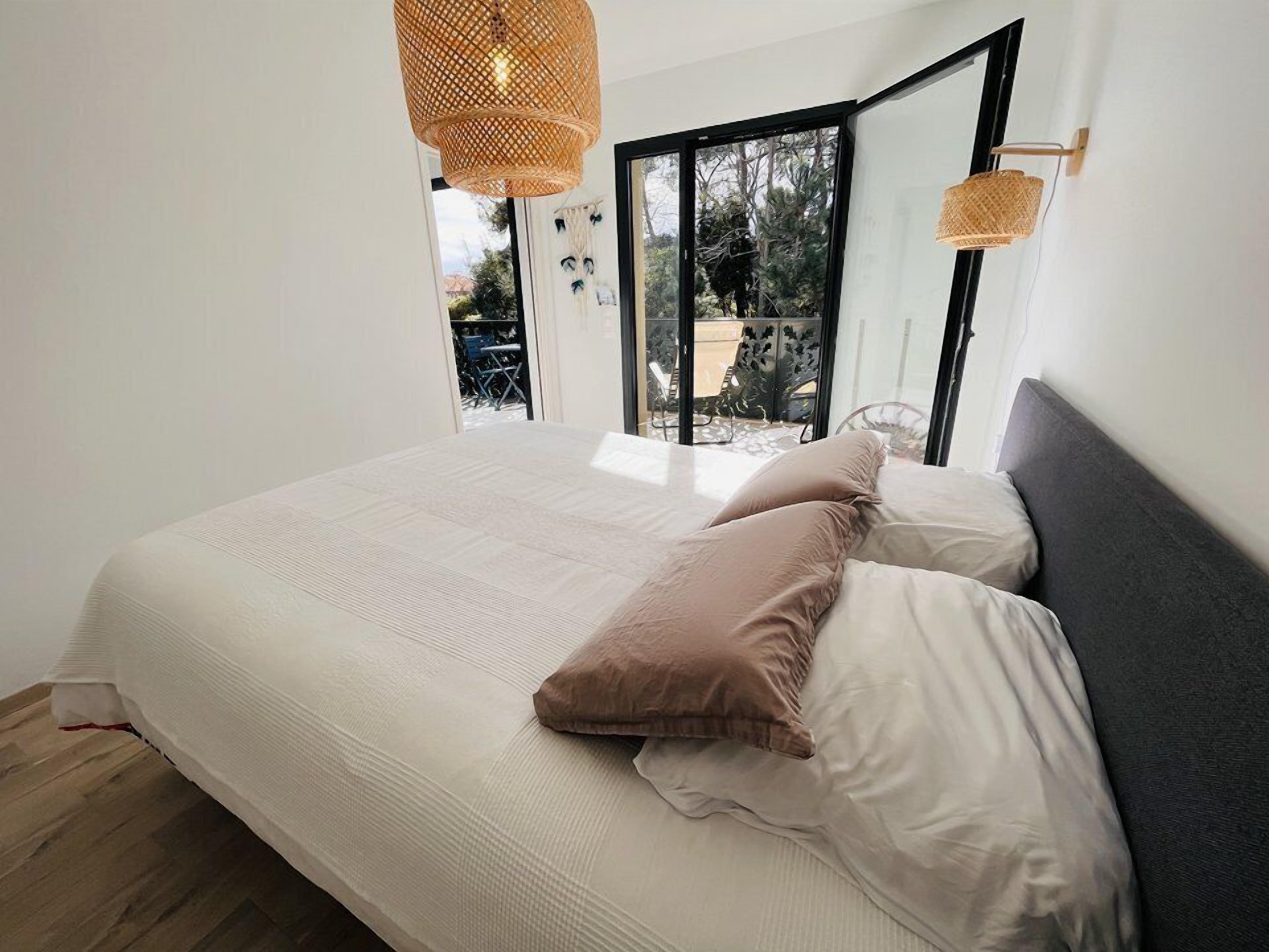 A bedroom with a large bed, a window with a view of outdoor furniture, and a woven light fixture.