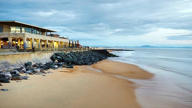 Un hotel frente a la playa con una larga costa rocosa y un edificio con grandes ventanales con vistas al mar.