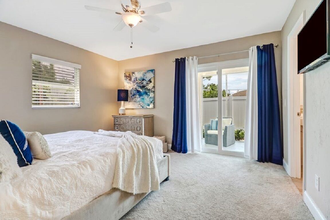 Une chambre à coucher comprenant un lit, un ventilateur de plafond, une commode, une fenêtre avec des stores et une vue sur l’extérieur, visible à travers une porte vitrée.