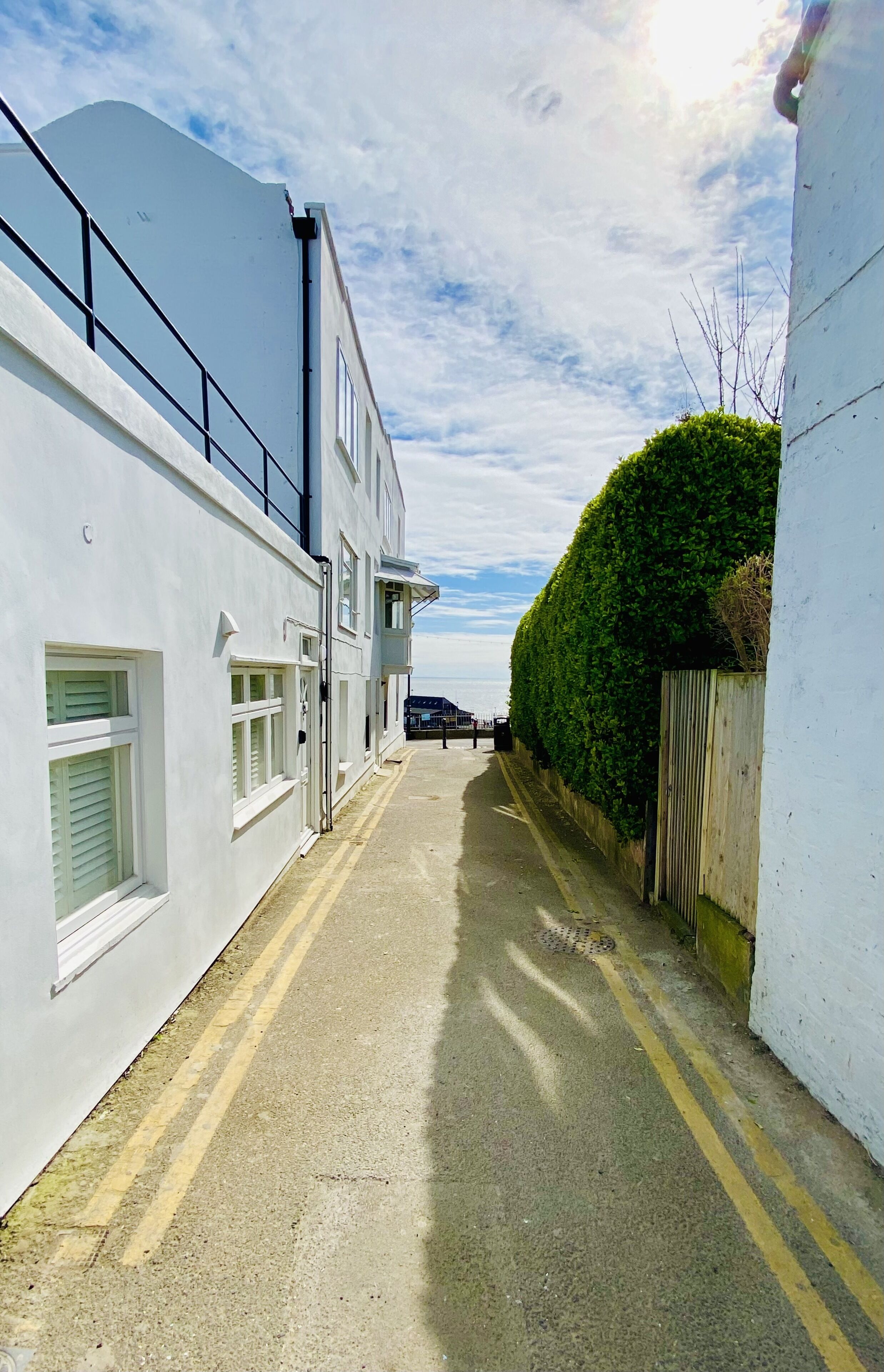 A narrow street with a white building on the left and a green hedge on the right.