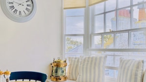 A dining area with a wooden table, chairs, a clock, and a window seat with cushions. - Nickleby Nook By the Sea - Meters to the Beach! (Broadstairs)
