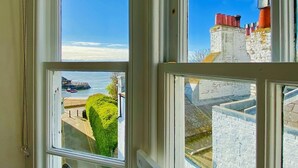 Quarto com uma grande janela com vista para um porto, um edifício de pedra com chaminé vermelha e um conjunto de sebes bem aparadas.