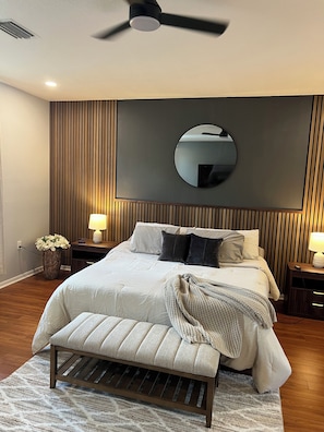 Um quarto com cama, mesas de cabeceira, um espelho redondo, um ventilador de teto e um banco.