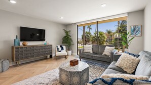 A modern living room with a sofa, a coffee table, a TV, and a large window with a view of palm trees. - Updated 3-Bedroom Getaway in Coronado Village (Coronado)
