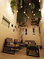 Una sala de estar acogedora con un conjunto de sofás, una mesita de centro y plantas colgantes. Hay dos ventanas y una puerta.