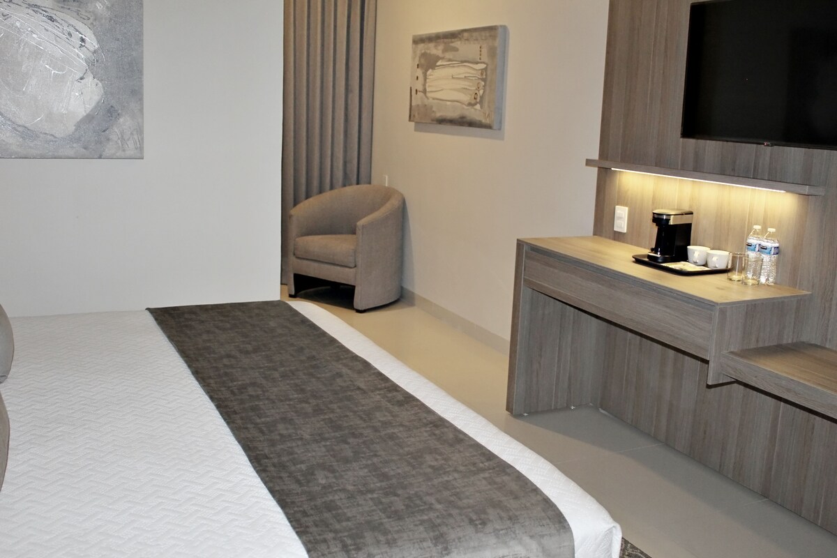 A modern hotel room with a bed, a desk with a coffee maker, and a TV on the wall.