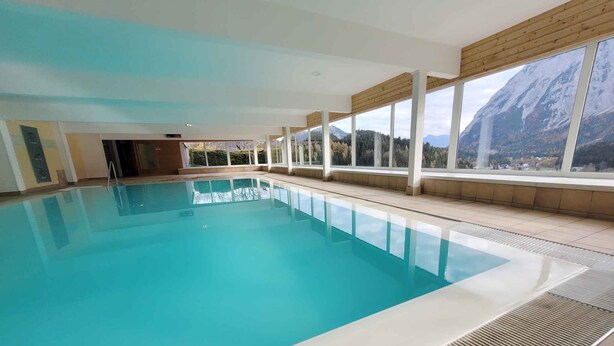 Ein großer Innenpool mit klarem, blauem Wasser, umgeben von großen Fenstern mit Blick auf die Berge.
