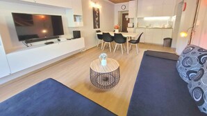 Ein modernes Wohnzimmer mit Flachbildfernseher, einem Essbereich mit Tisch und Stühlen, einer Küche mit weißen Schränken und einem gemusterten Sofa.