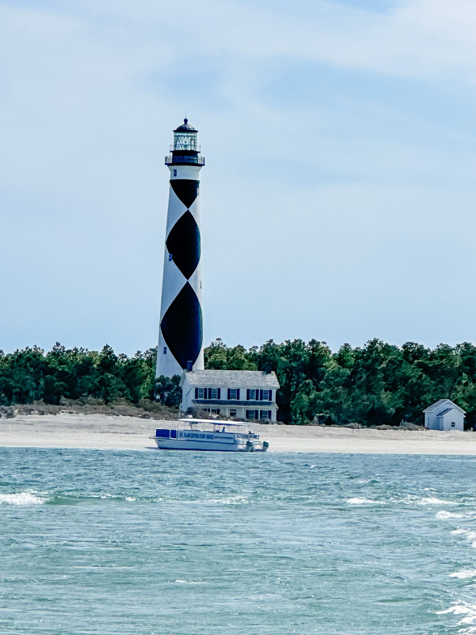 Un phare au design unique, avec des bandes noires et blanches en spirale, une maison de plage en arrière-plan et un bateau sur l’eau.