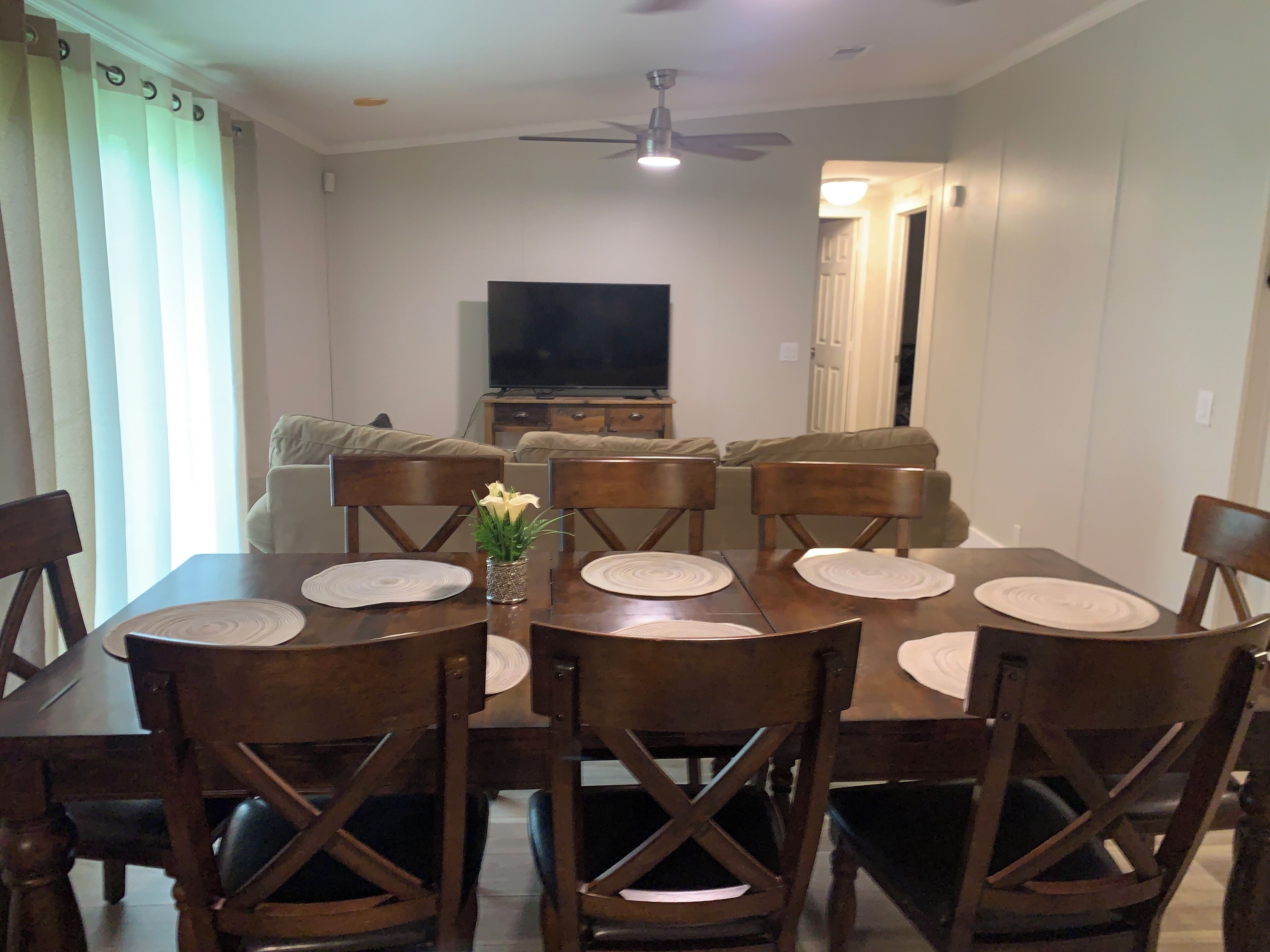 Un comedor con mesa y sillas de madera, un florero con flores y un televisor al fondo.