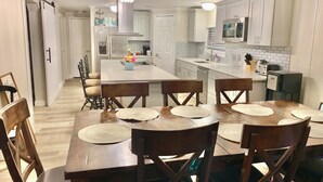 A modern kitchen with white cabinets, a central island, and a dining area with wooden chairs and placemats. - Cozy 5 Bedroom 85” TV Home in a Secluded Area (Jacksonville)