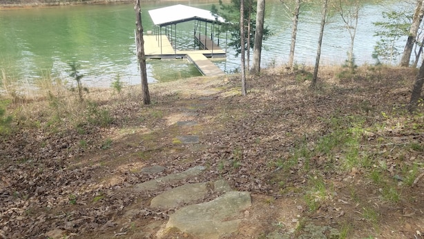 A stone pathway leading to a dock over a calm lake, surrounded by trees and a covered seating area.