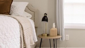 A bedroom with a bed, a bedside table with a lamp, a painting on the wall, and a window with curtains.
