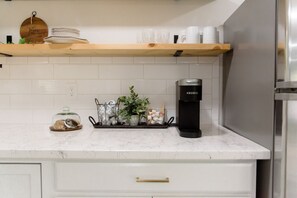 Una cocina con encimeras de mármol blanco, una cafetera Keurig y una estantería de madera con copas y platos.