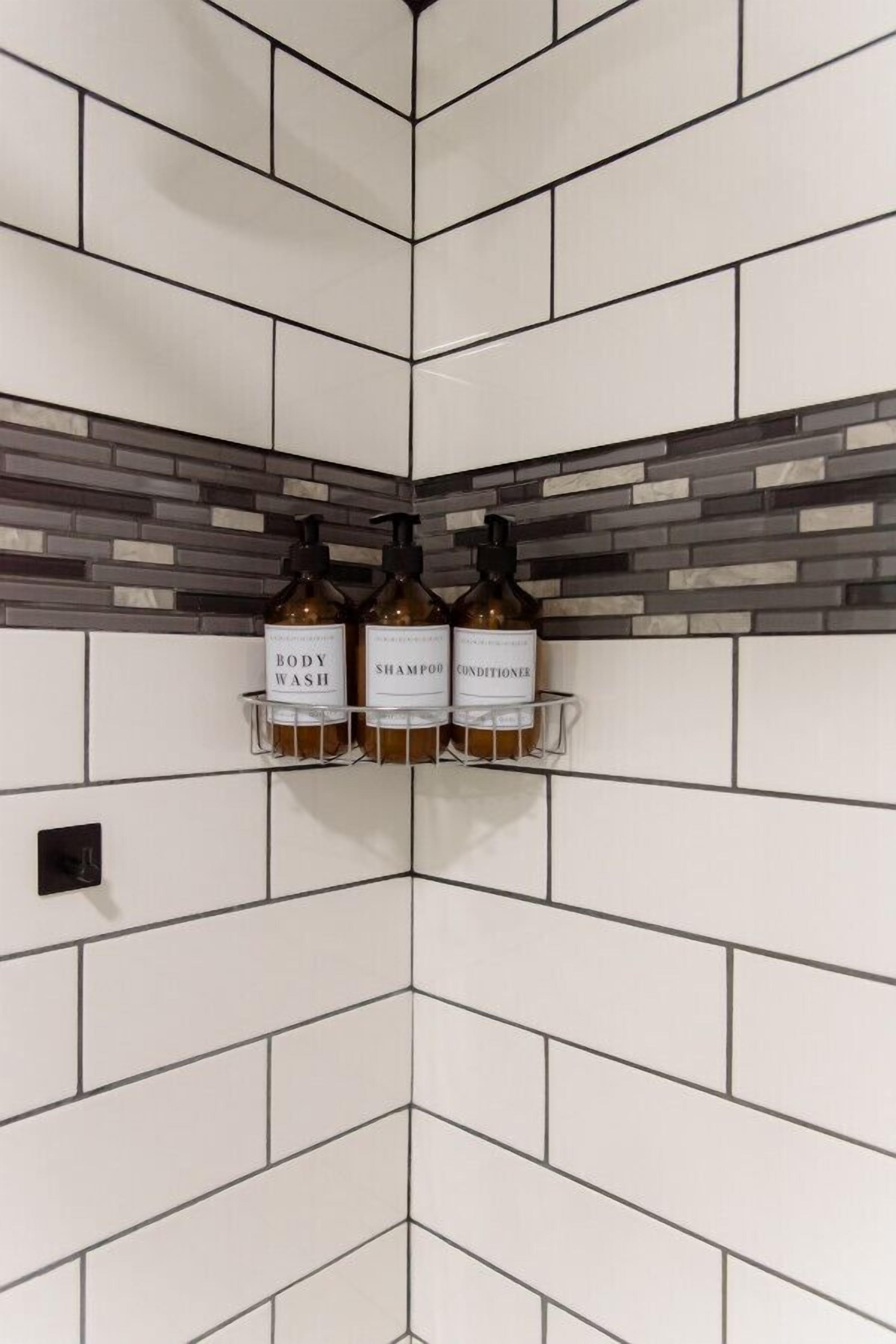 Een badkamer met witte metrotegels en een accentmuur met grijs en wit mozaïek. Op een plank staan drie flessen douchegel, shampoo en conditioner.