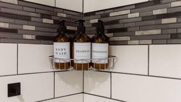 Een badkamer met witte metrotegels en een accentmuur met grijs en wit mozaïek. Op een plank staan drie flessen douchegel, shampoo en conditioner.
