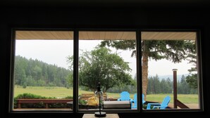 A room with a large window overlooking a grassy area with trees and a wooden deck with blue chairs. - Country Living in Kalispell Montana (Kalispell)