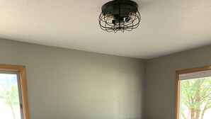 A bedroom with a large bed, two bedside tables with lamps, a ceiling light fixture, and windows with a view of trees. - Country Living in Kalispell Montana (Kalispell)