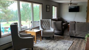 A spacious living room with a large window, a ceiling fan, and a flat-screen TV mounted on the wall. - Country Living in Kalispell Montana (Kalispell)