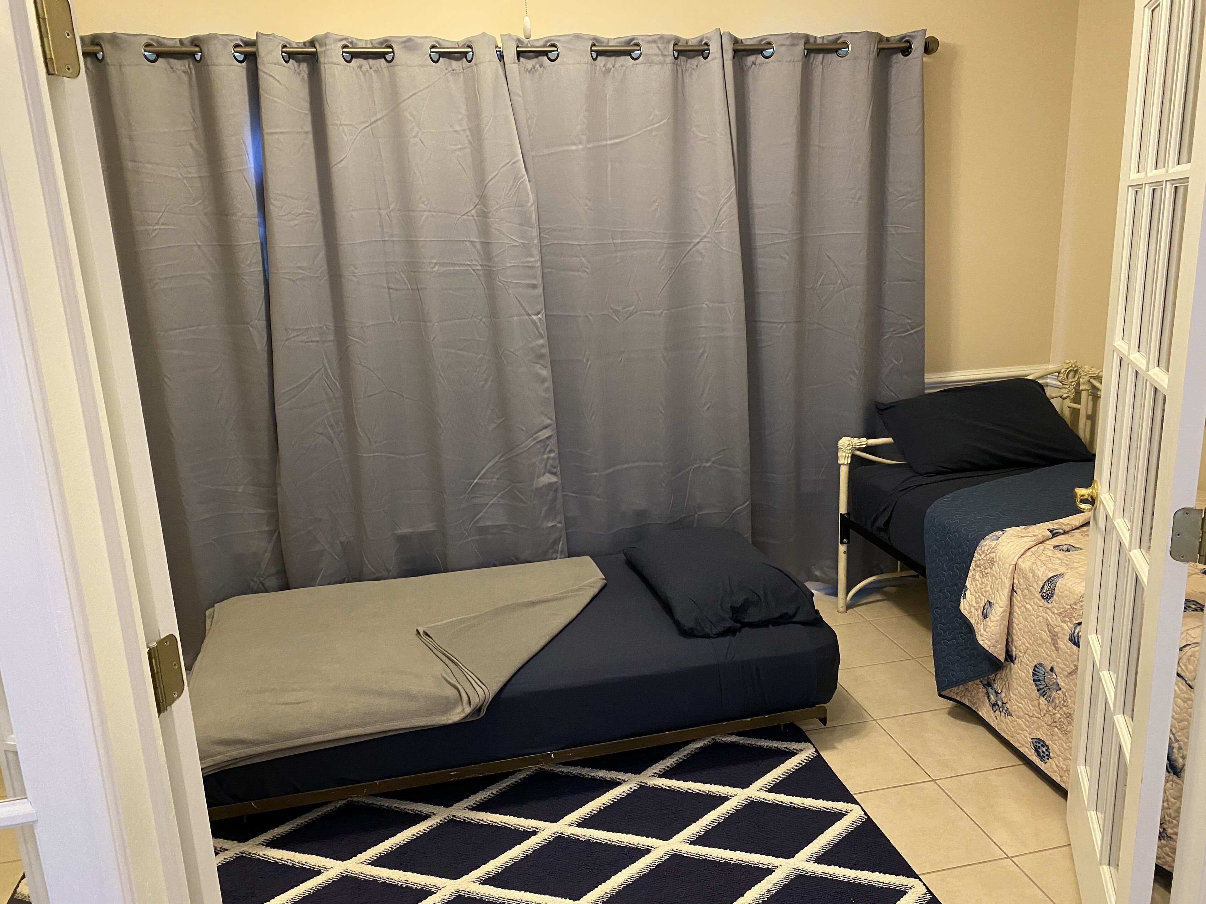 A small, compact bedroom with a single bed, a second smaller bed, a window with curtains, and a patterned rug.