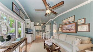 A living room with a sofa, coffee table, and ceiling fan. There are framed pictures on the wall and a kitchen in the background.
