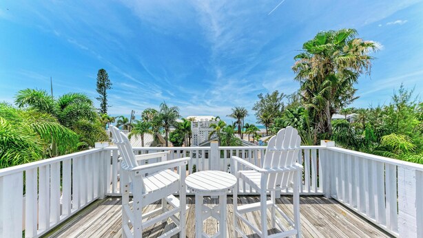 A wooden deck with white chairs and a table, surrounded by lush greenery and a clear blue sky.