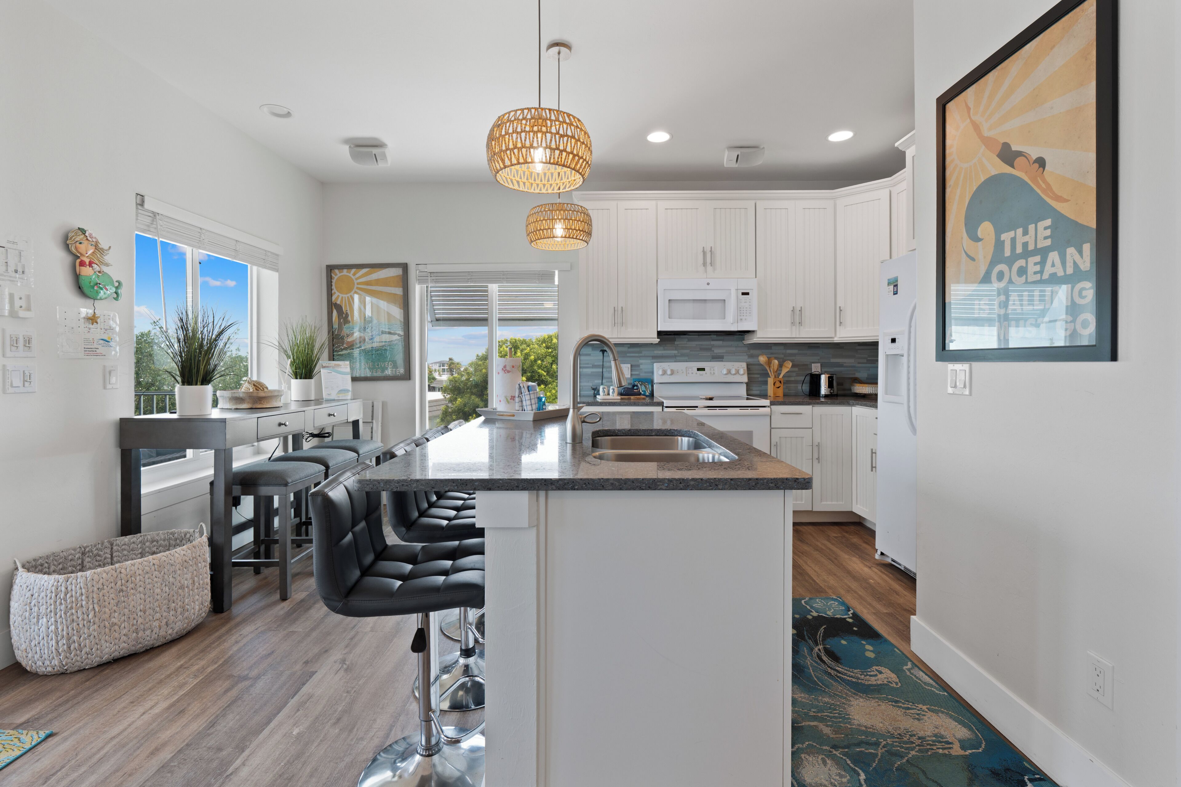A modern kitchen with white cabinets, a central island with bar stools, a large window, and a hanging pendant light.