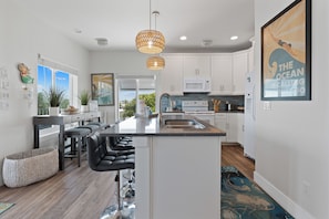 A modern kitchen with white cabinets, a central island with bar stools, a large window, and a hanging pendant light. - Bluewater Vista is a super cool Mid Island Pool Home with Hot Tub (Fort Myers Beach)