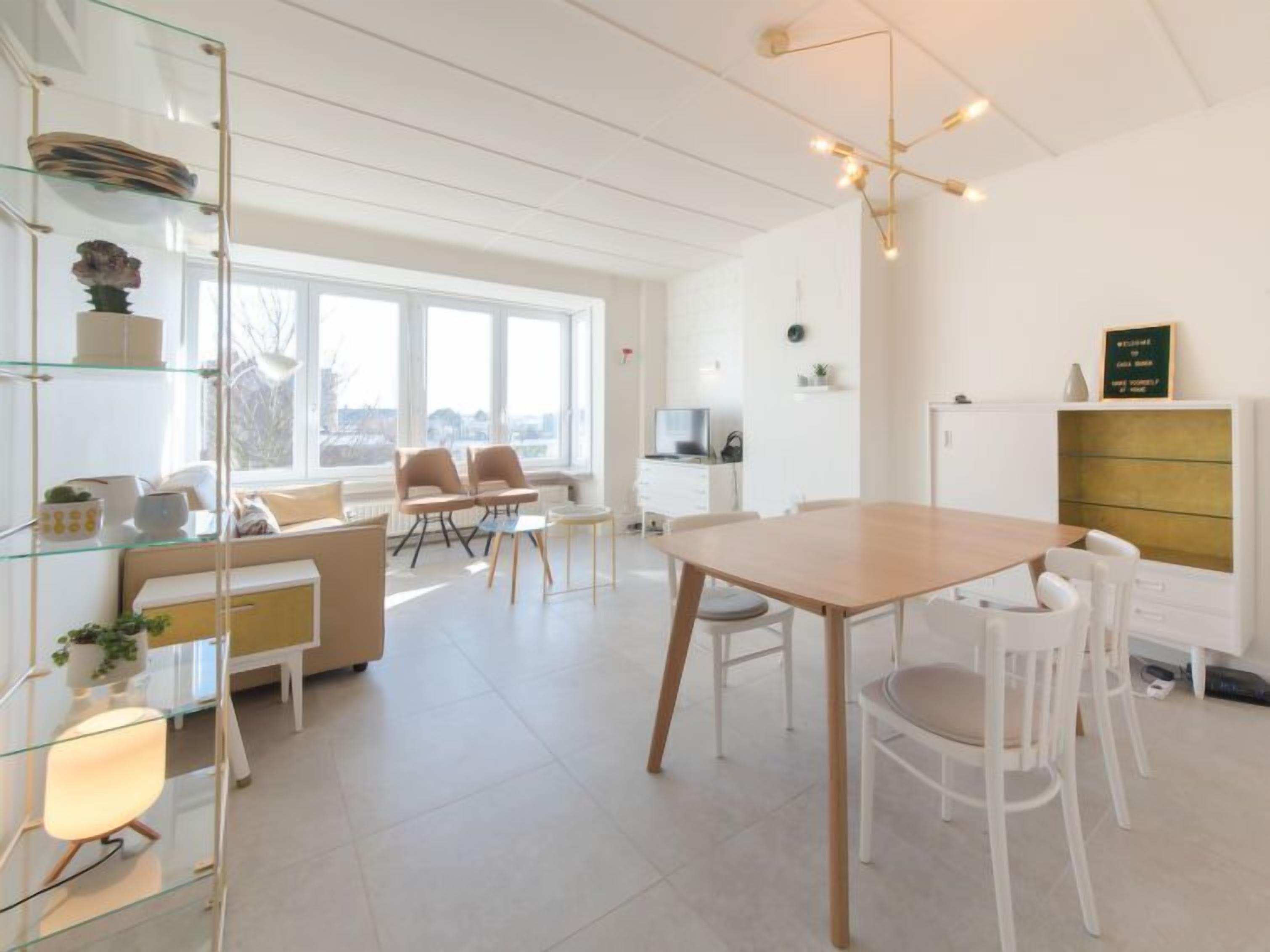 A modern, well-lit living room with a dining area, large windows, and a minimalist design.