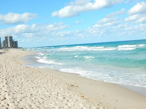 A sandy beach with waves, a clear sky, and buildings in the background.