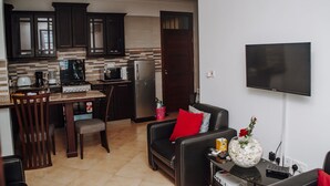 A modern kitchen with dark cabinets, a dining area with a table and chairs, and a living area with a sofa and a flat-screen TV.