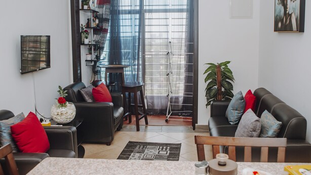 A modern living room with a black leather sofa, a coffee table, and a dining table. There is a large window with sheer curtains, a potted plant, and a painting on the wall.